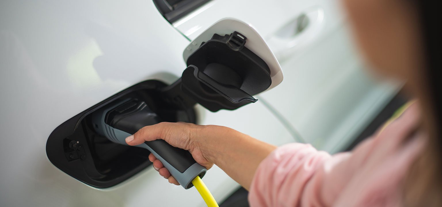Frau steckt ein Ladekabel in die Buchse eines weissen Elektroautos.