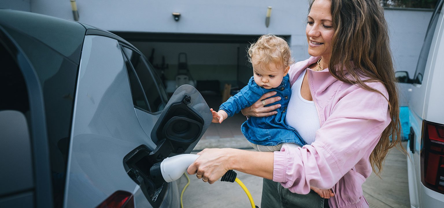 Mutter mit Kleinkind lädt ein Elektroauto vor der Garage auf.