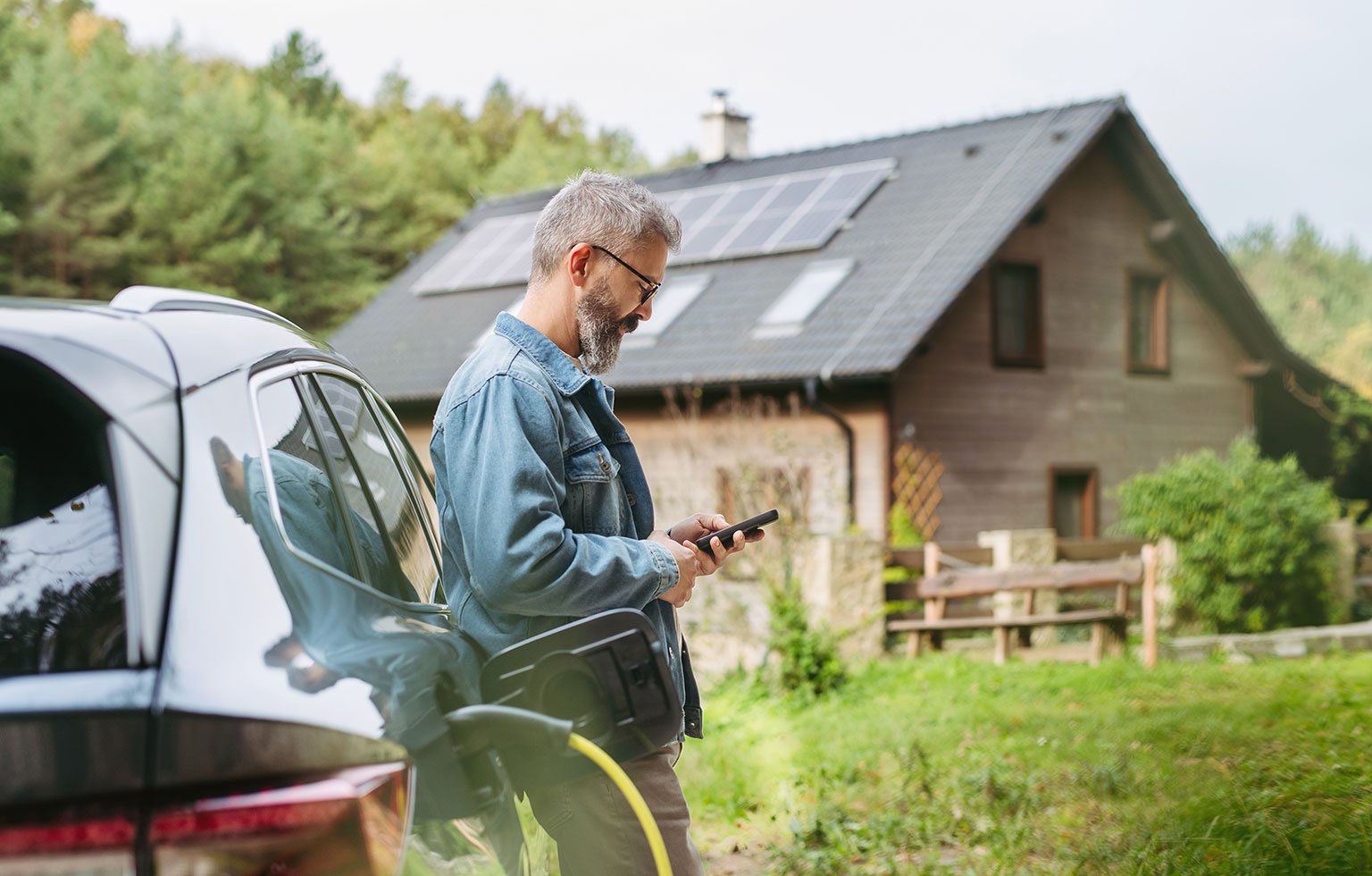 Mann steht an einem E-Auto mit Ladekabel und nutzt sein Smartphone vor einem Haus mit Solaranlage.