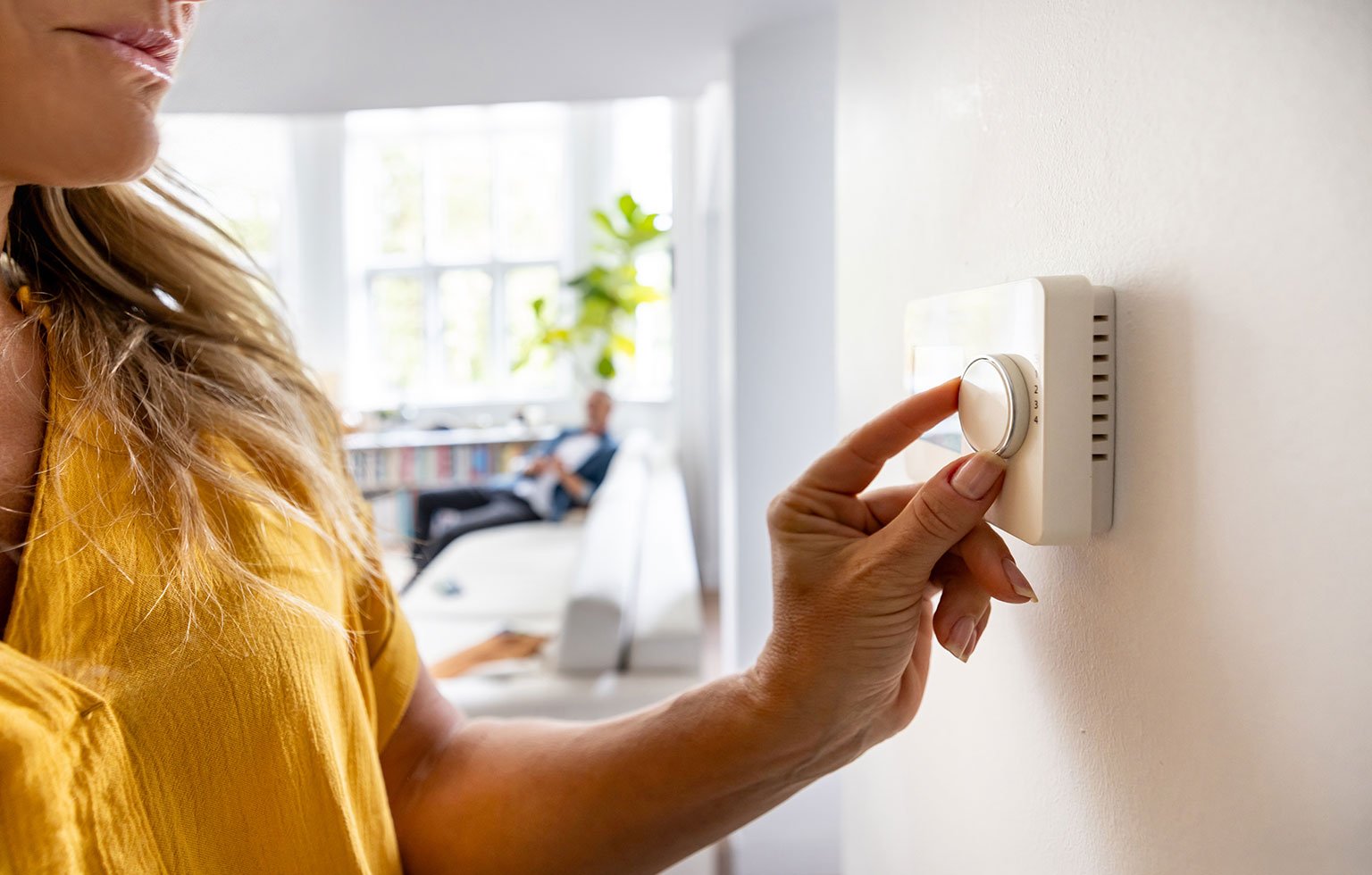 Frau regelt die Temperatur an einem Wandthermostat in einem hellen Wohnzimmer.