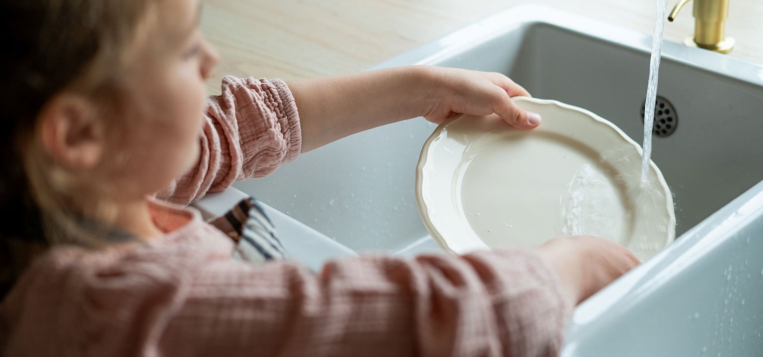 Ein Kind spült einen Teller unter fliessendem Wasser am Spülbecken.
