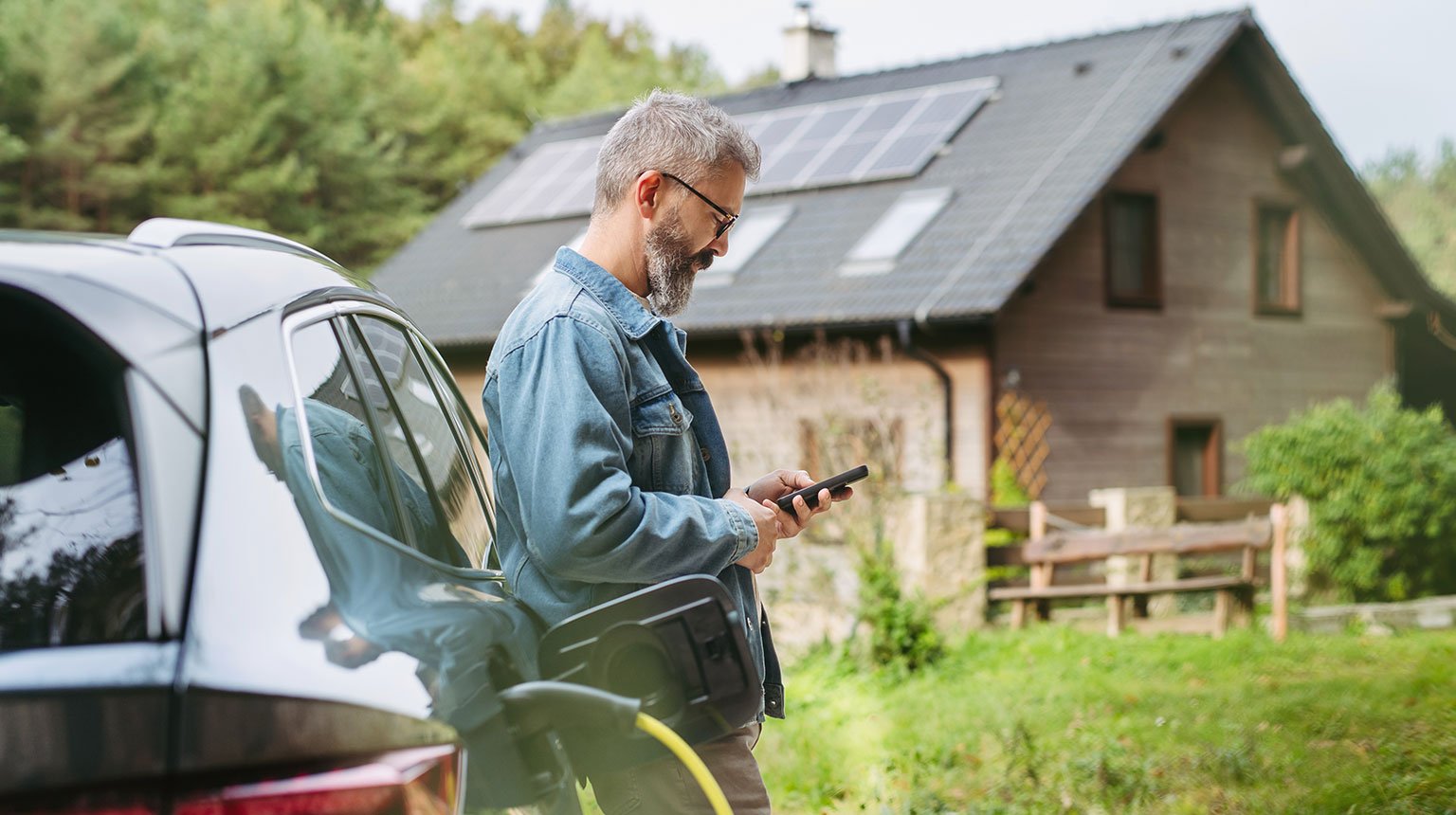Ein Mann lehnt an einem Elektroauto beim Laden und schaut auf sein Smartphone; im Hintergrund ein Haus mit Solarpanels auf dem Dach.