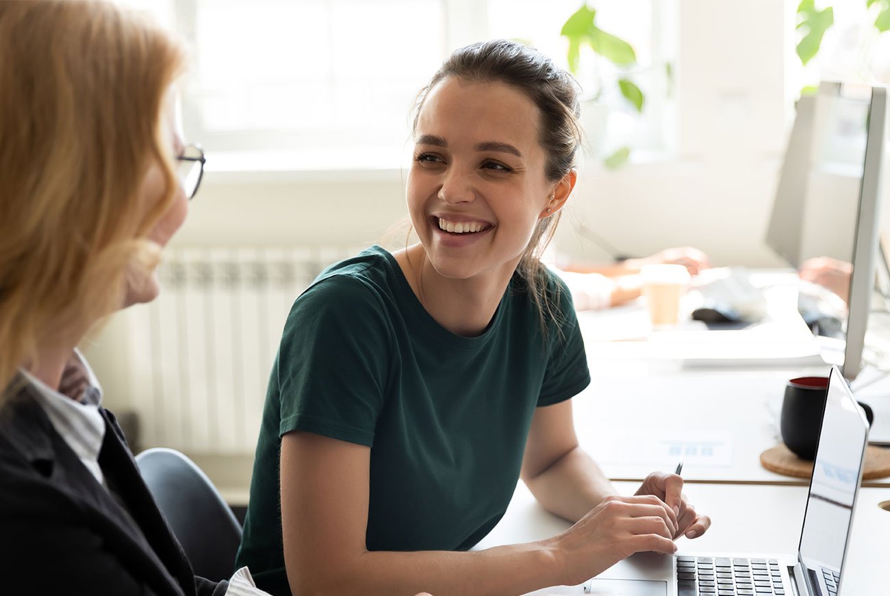 Zwei Frauen unterhalten sich an einem Schreibtisch mit Laptop im Büro.