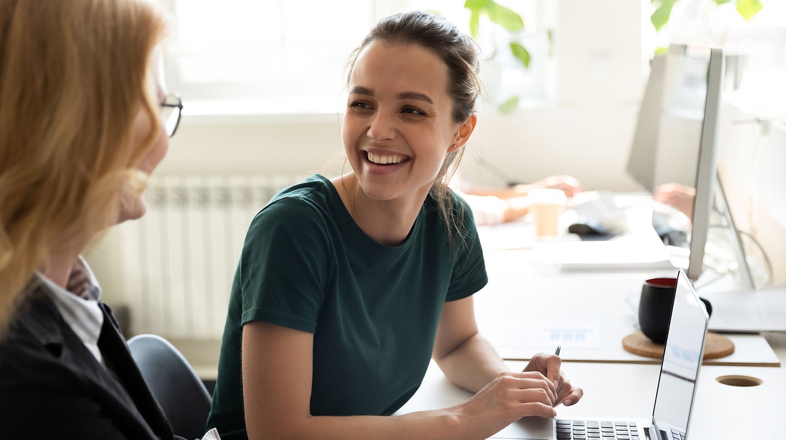 Zwei Frauen unterhalten sich an einem Schreibtisch mit Laptop im Büro.