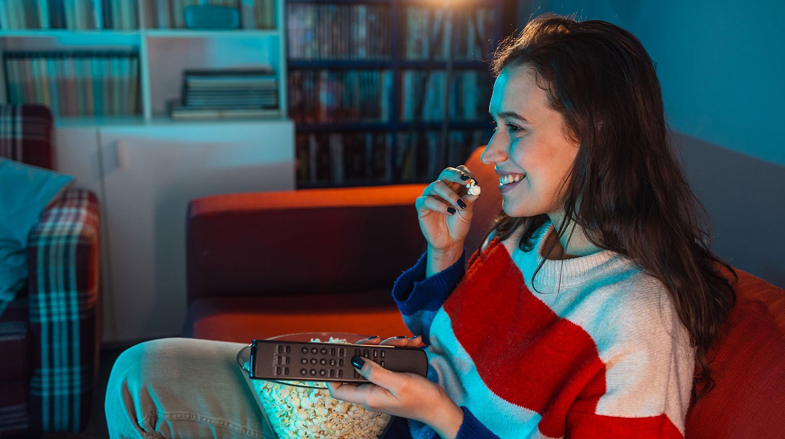 Junge Frau sitzt auf dem Sofa, isst Popcorn und schaut fern mit Fernbedienung in der Hand.