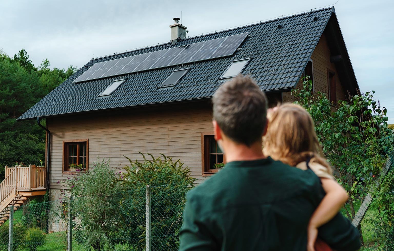 Vater mit Kind blickt auf ein Holzhaus mit Solarpanels auf dem Dach.