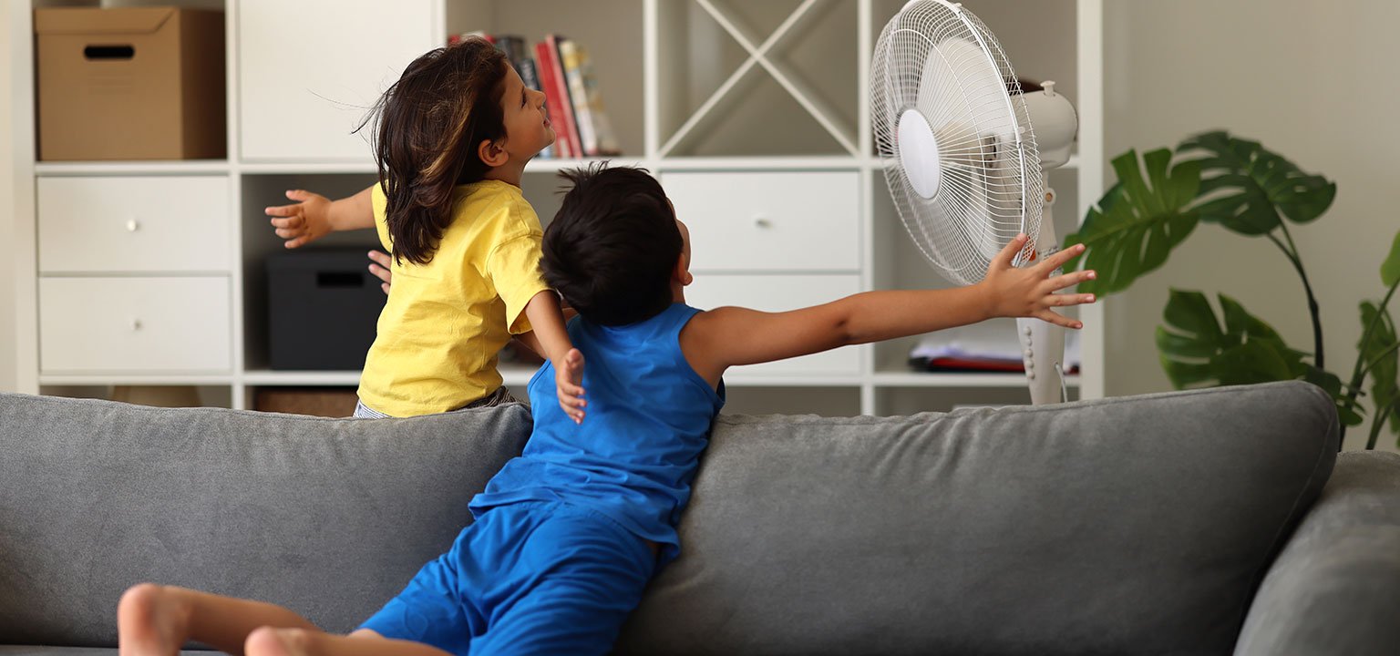 Zwei Kinder geniessen die Abkühlung vor einem Ventilator im Wohnzimmer.