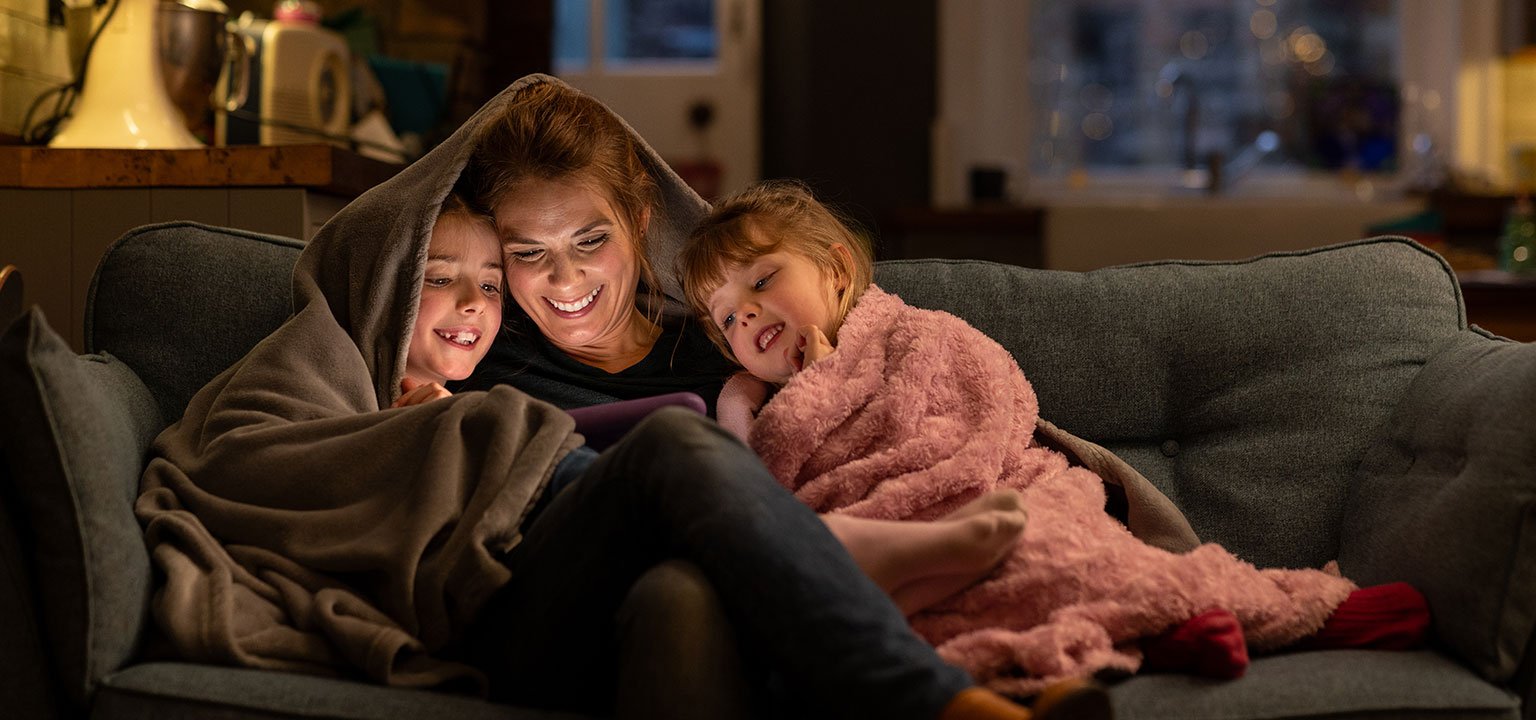 Mutter sitzt mit zwei Kindern unter Decken auf dem Sofa und schaut mit ihnen ein Tablet.