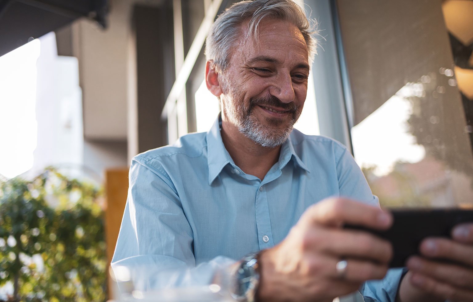 Mann mittleren Alters sitzt draussen und schaut lächelnd auf sein Smartphone.