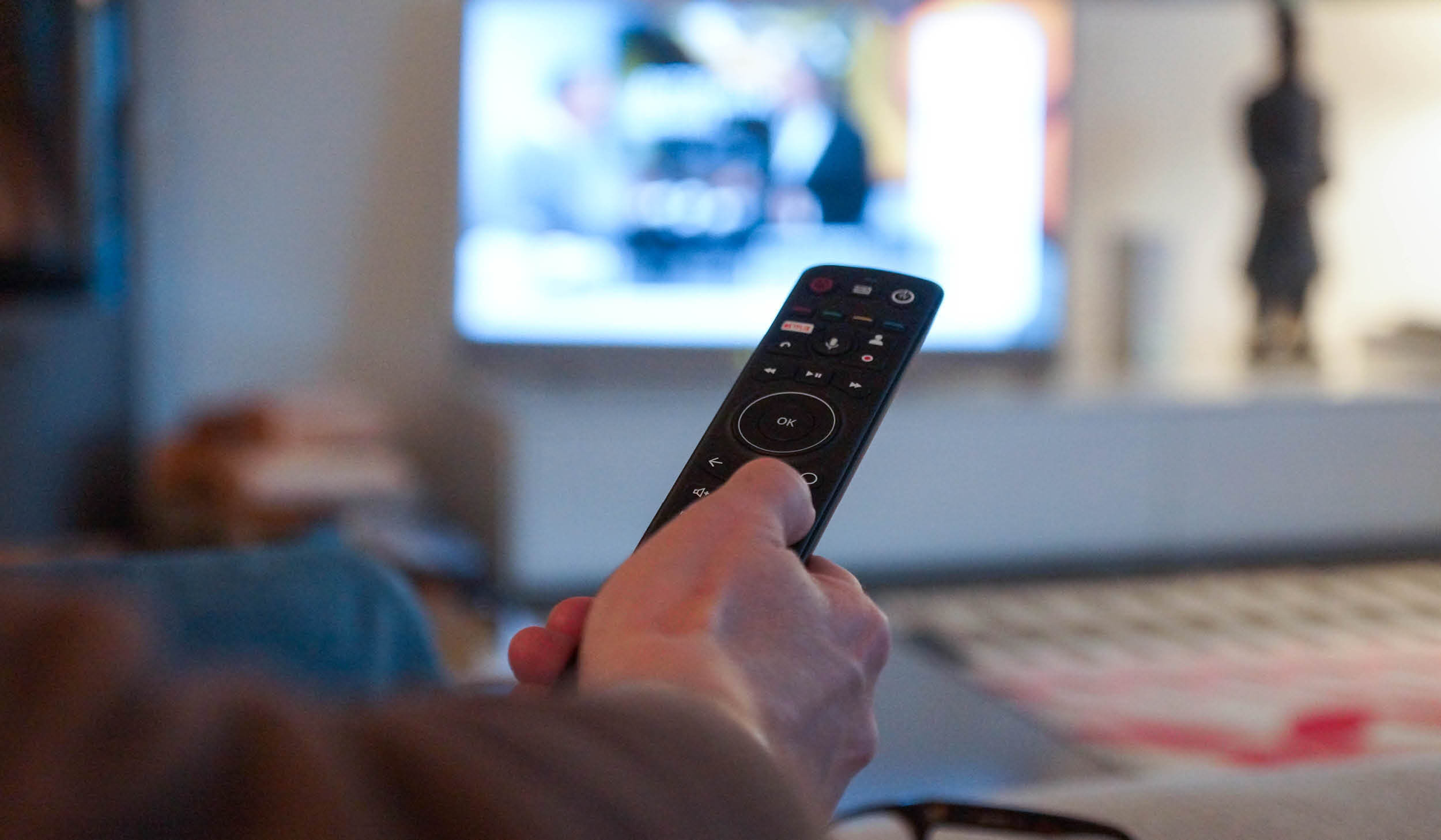 Person mit Fernbedienung vor einem eingeschalteten Fernseher.