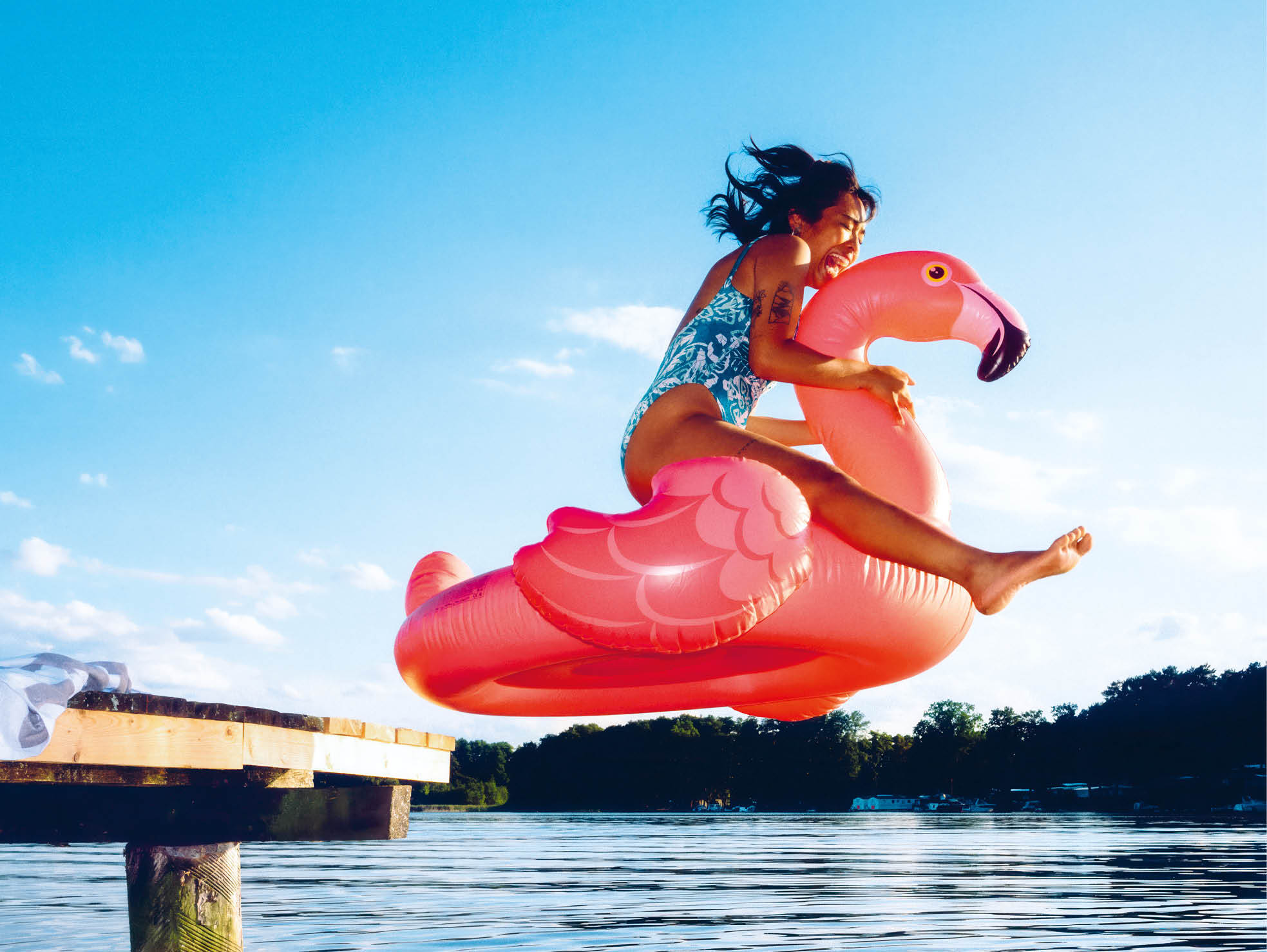 Junge Frau springt mit aufblasbarem Flamingo in den See.