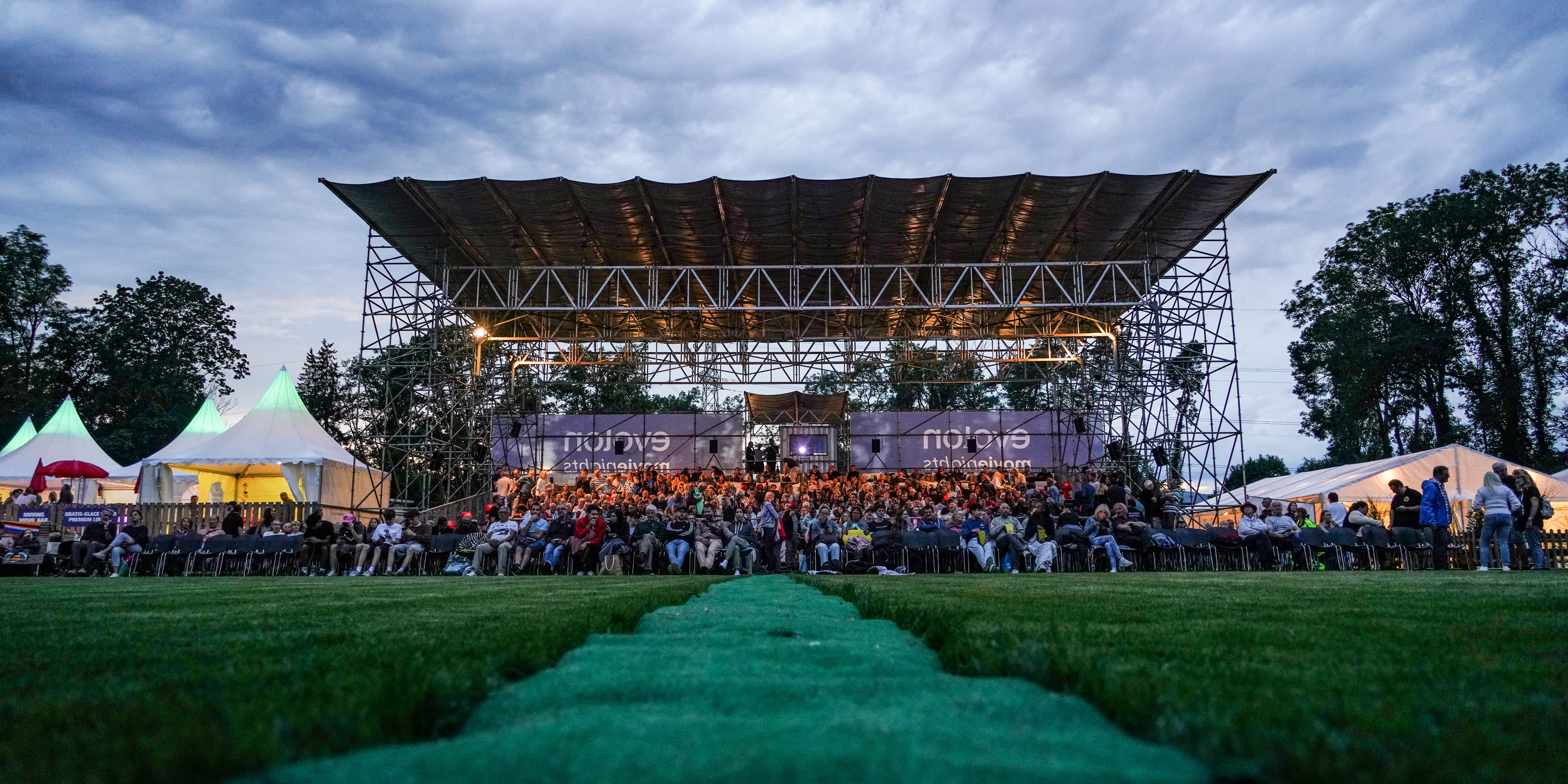 Open-Air-Bühne mit großem Publikum auf einer Wiese bei Abenddämmerung unter bewölktem Himmel.