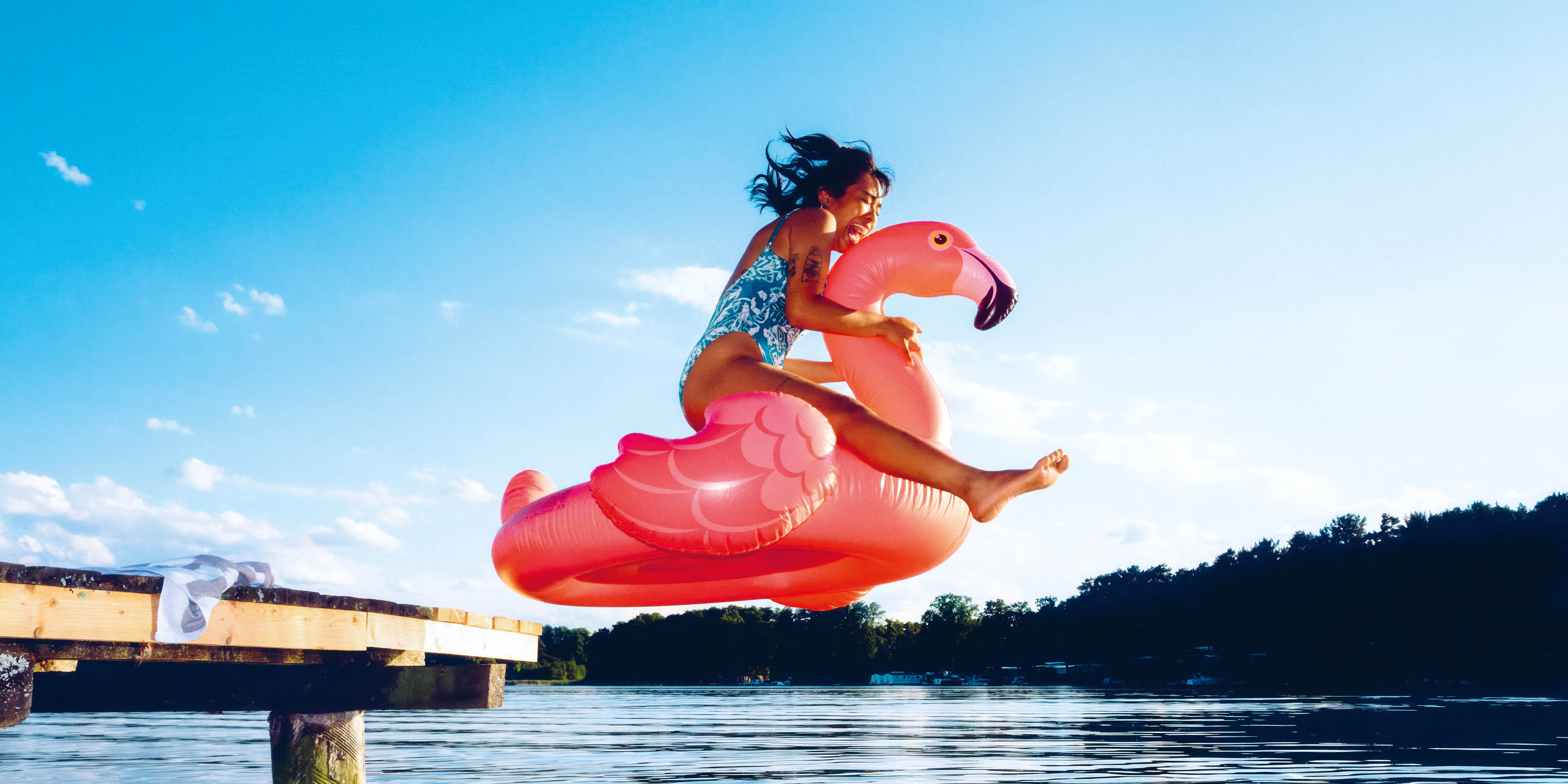 Person springt mit einem pinken Flamingo-Schwimmring von einem Steg in einen Badesee.