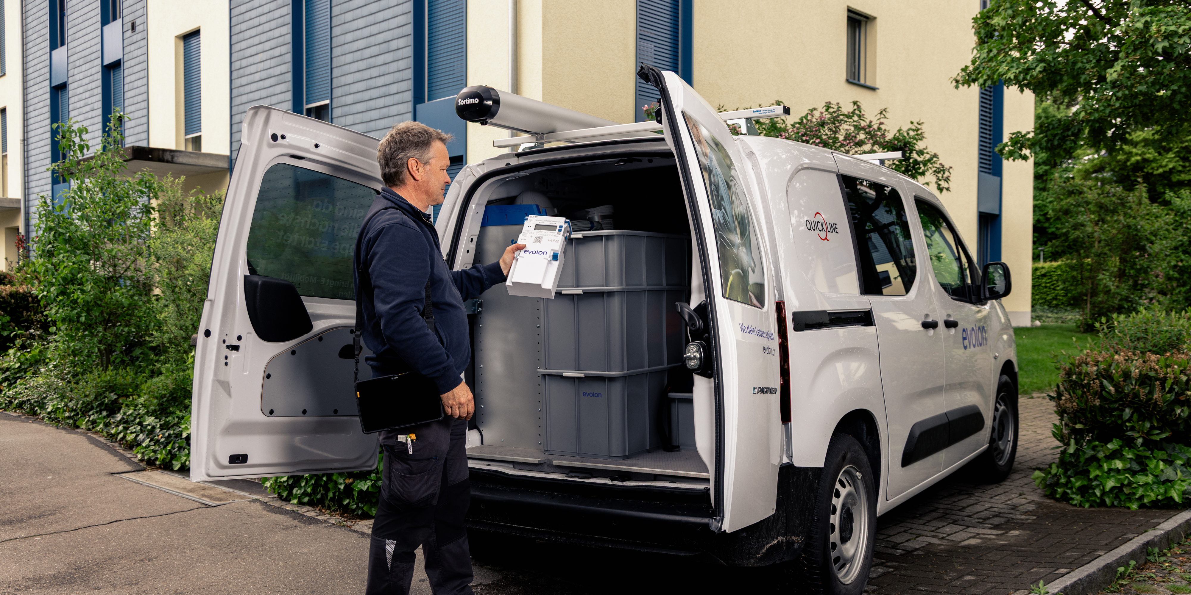 Person steht vor dem offenen Laderaum eines weissen Lieferwagens und hält ein Gerät in der Hand.