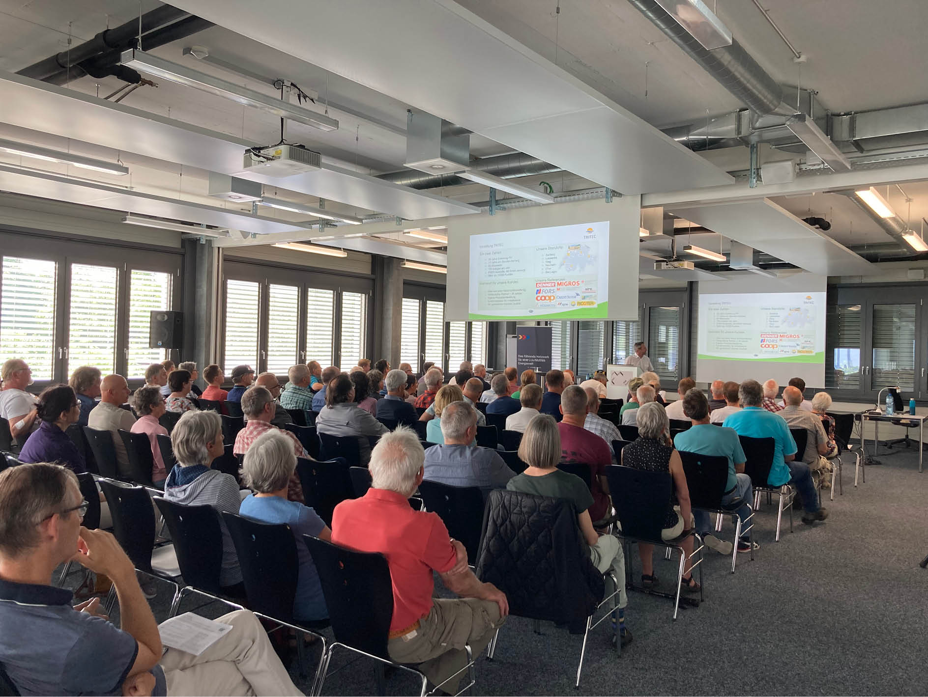 Grosser Konferenzraum mit sitzendem Publikum, das einem Vortragenden an der Leinwand mit Präsentation aufmerksam zuhört.