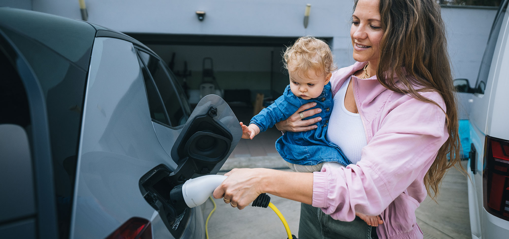 Mutter mit Kleinkind lädt ein Elektroauto vor der Garage auf.