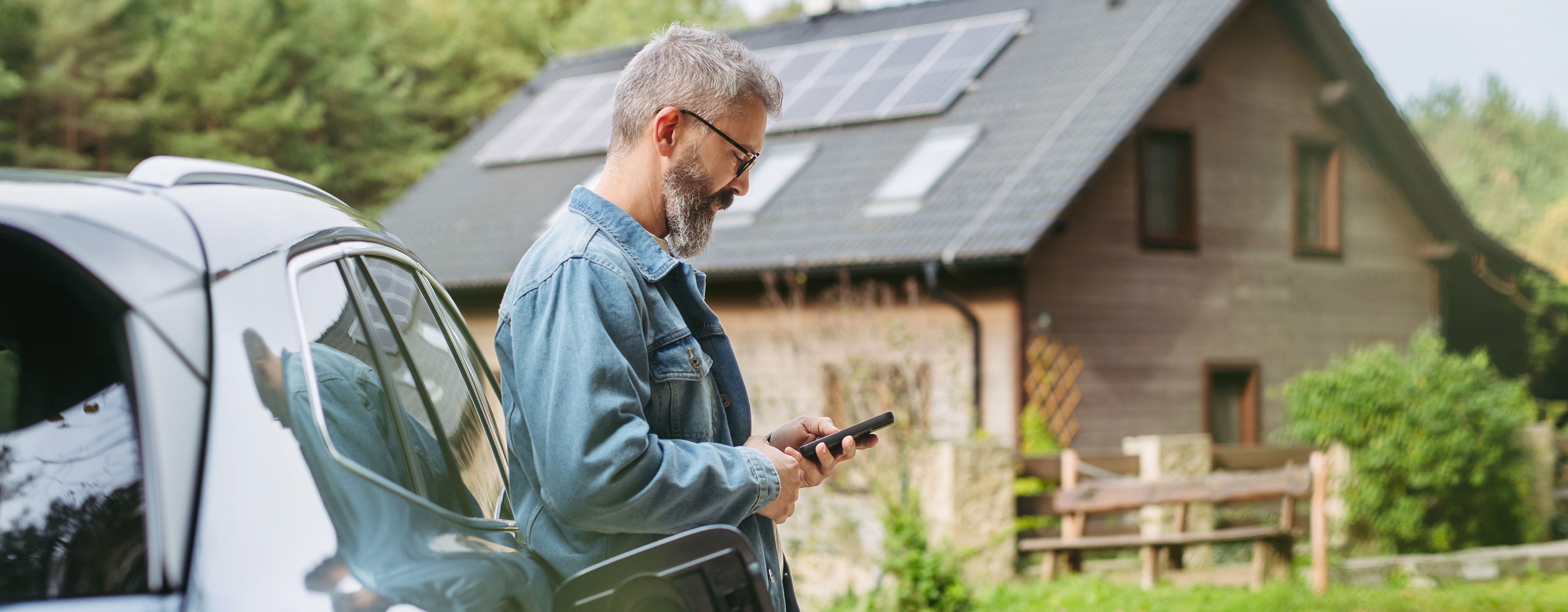 Mann steht an einem E-Auto mit Ladekabel und nutzt sein Smartphone vor einem Haus mit Solaranlage.
