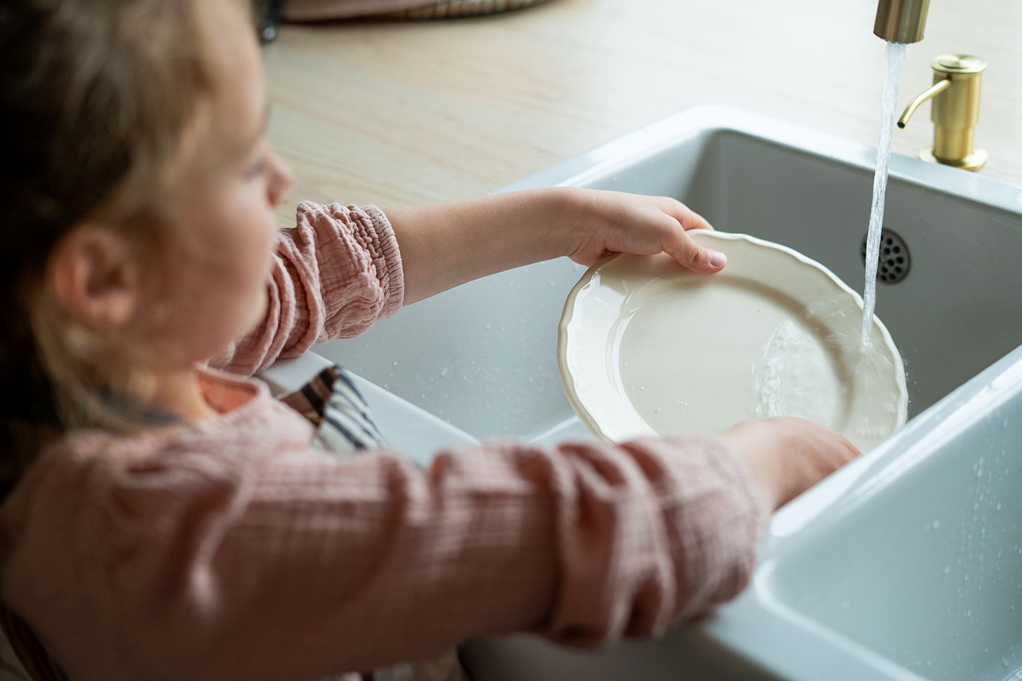 Ein Kind spült einen Teller unter fliessendem Wasser am Spülbecken.