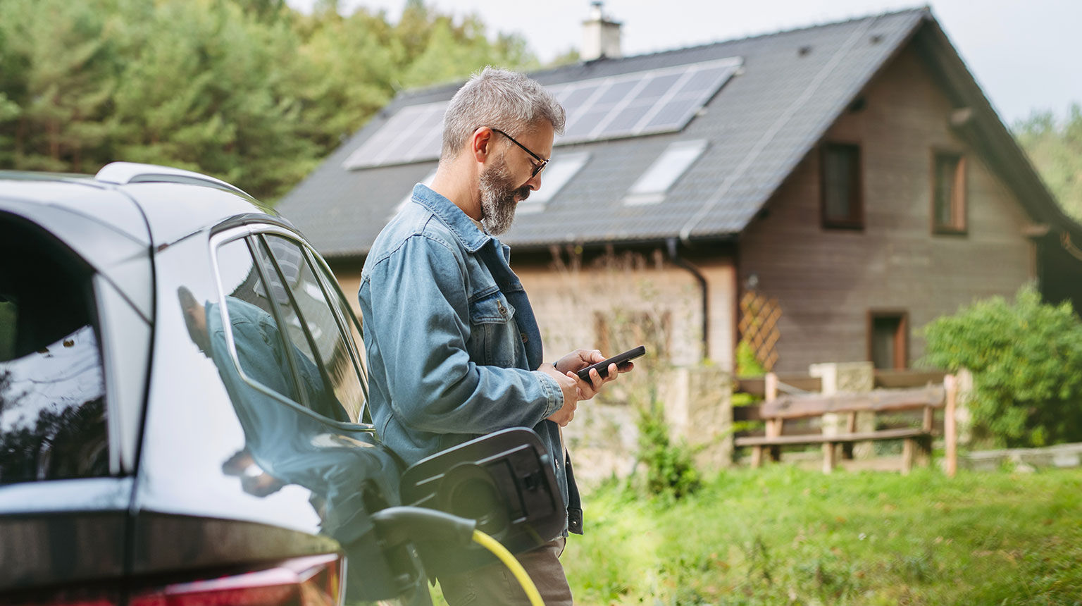 Ein Mann lehnt an einem Elektroauto beim Laden und schaut auf sein Smartphone; im Hintergrund ein Haus mit Solarpanels auf dem Dach.