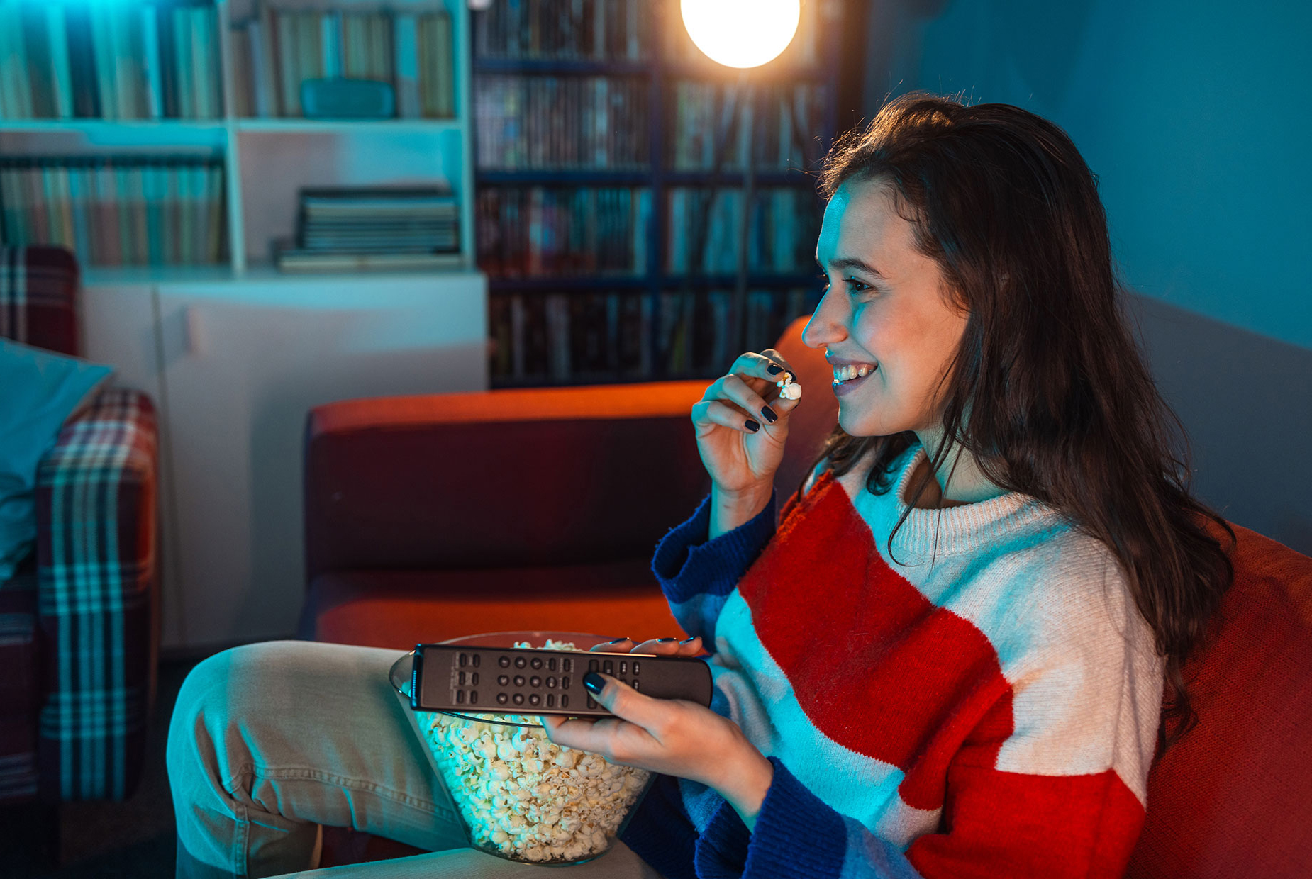Junge Frau sitzt auf dem Sofa, isst Popcorn und schaut fern mit Fernbedienung in der Hand.