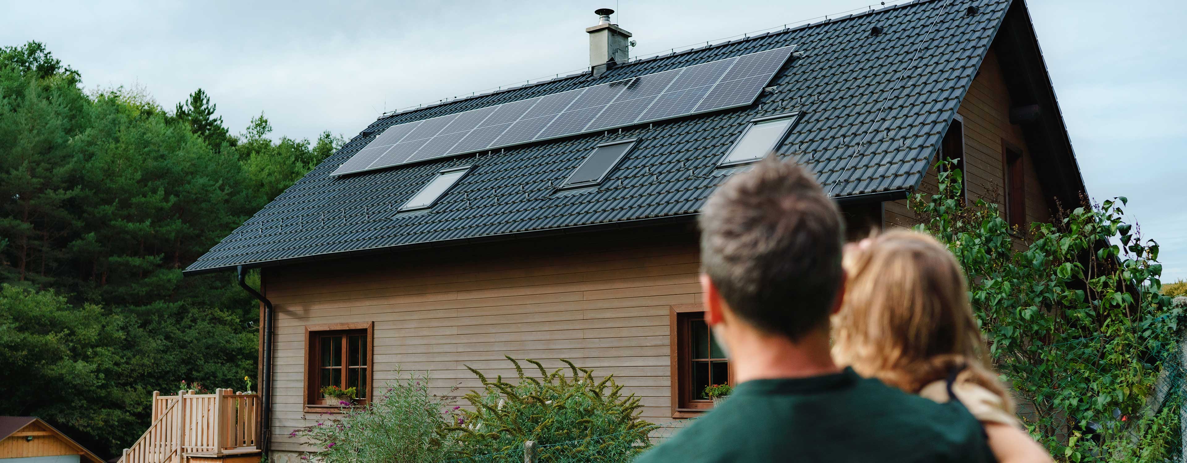 Vater mit Kind blickt auf ein Holzhaus mit Solarpanels auf dem Dach.