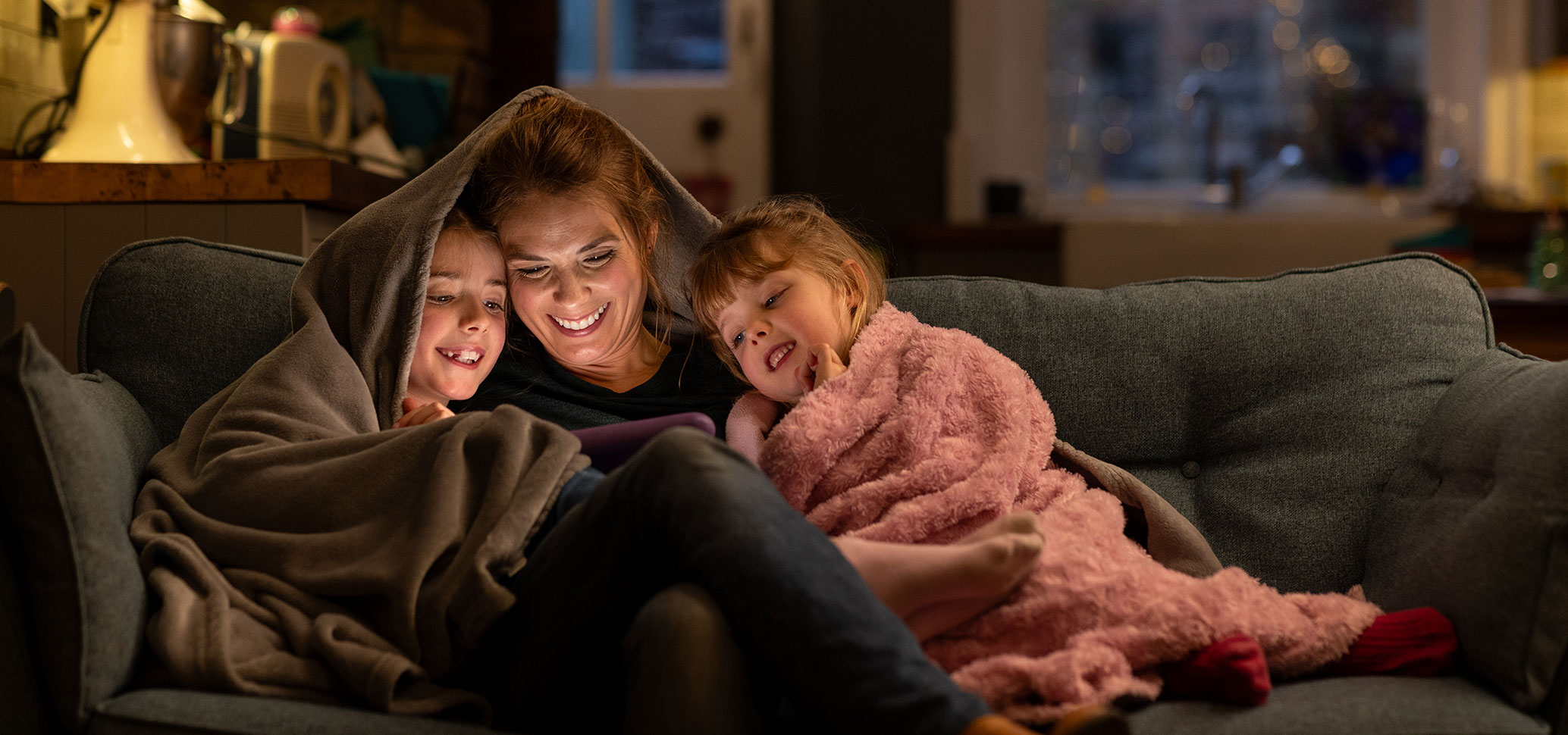Mutter sitzt mit zwei Kindern unter Decken auf dem Sofa und schaut mit ihnen ein Tablet.